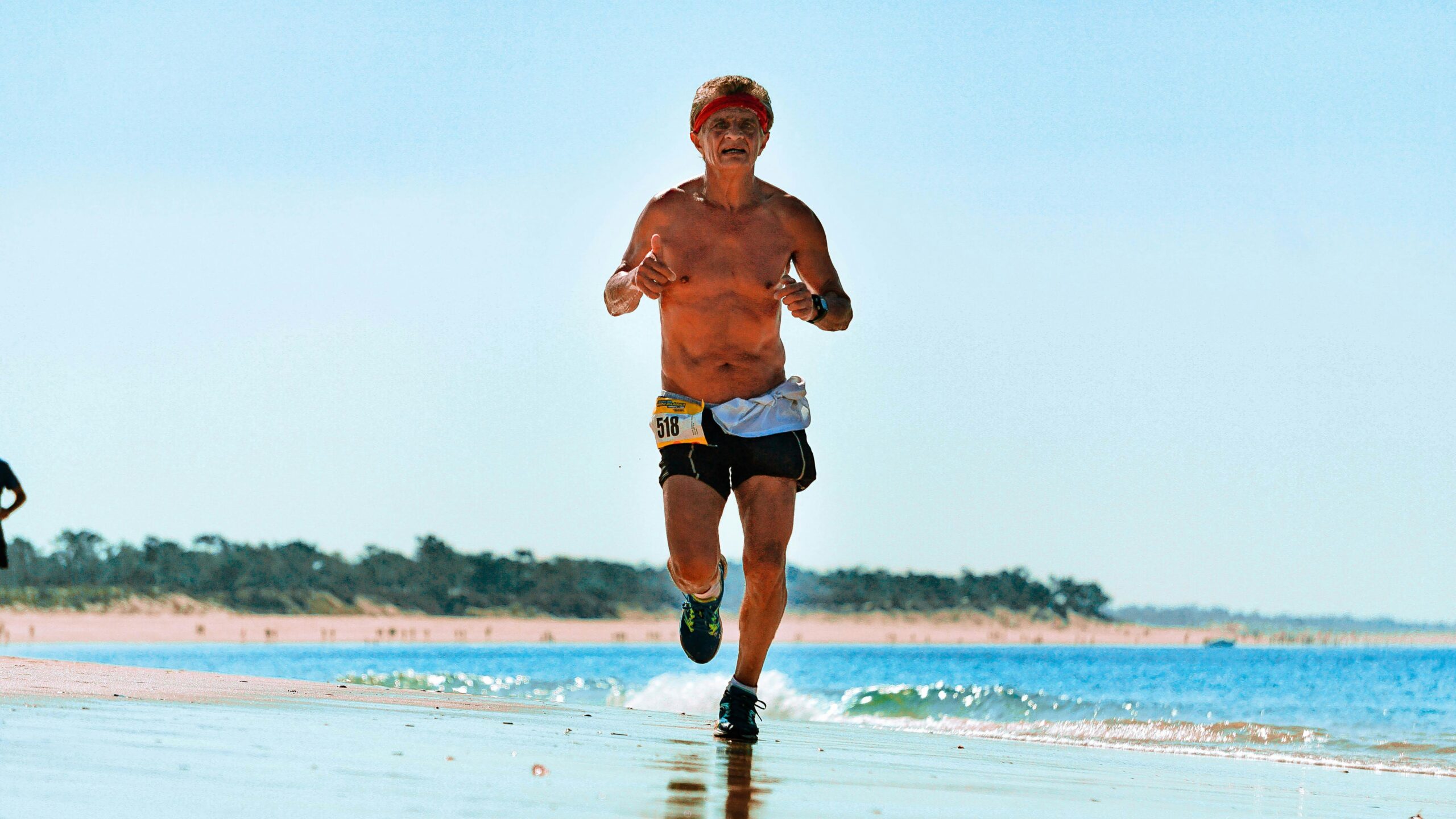 A senior man runs shirtless along a sunny beach, showcasing fitness and vitality.